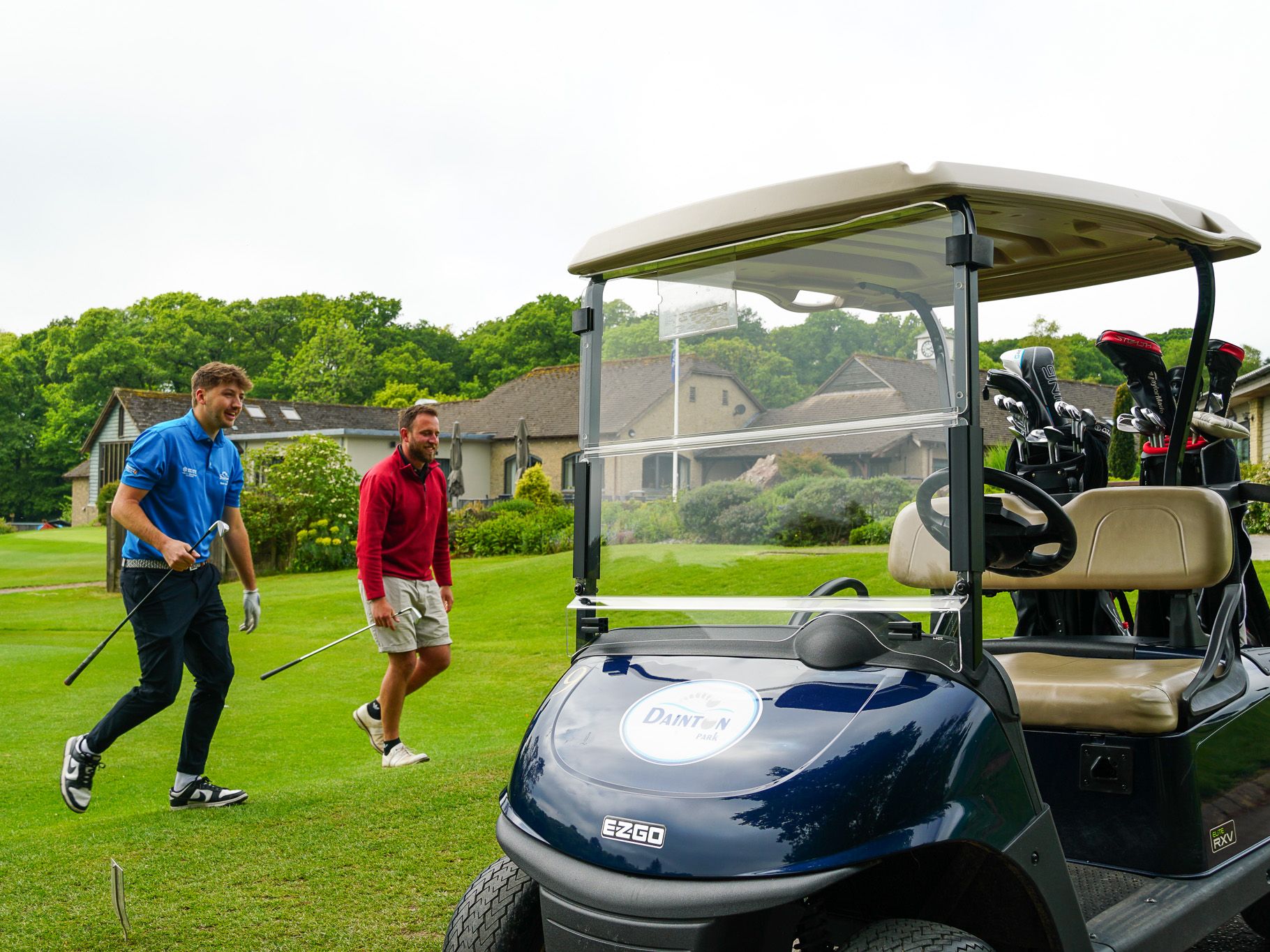 Two men walking on a golf course near a golf cart, holding golf clubs with a clubhouse and greenery in the background.