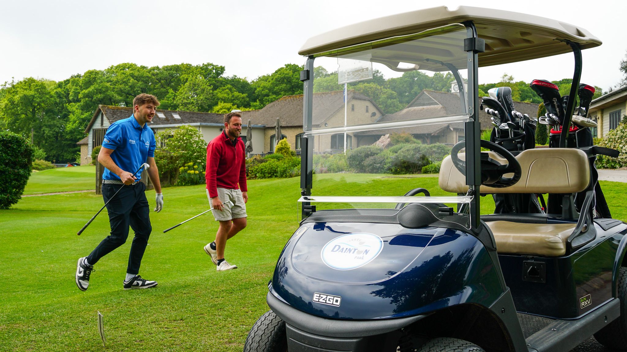 Two men walking on a golf course near a golf cart, holding golf clubs with a clubhouse and greenery in the background.