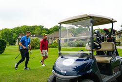 Two men walking on a golf course near a golf cart, holding golf clubs with a clubhouse and greenery in the background.