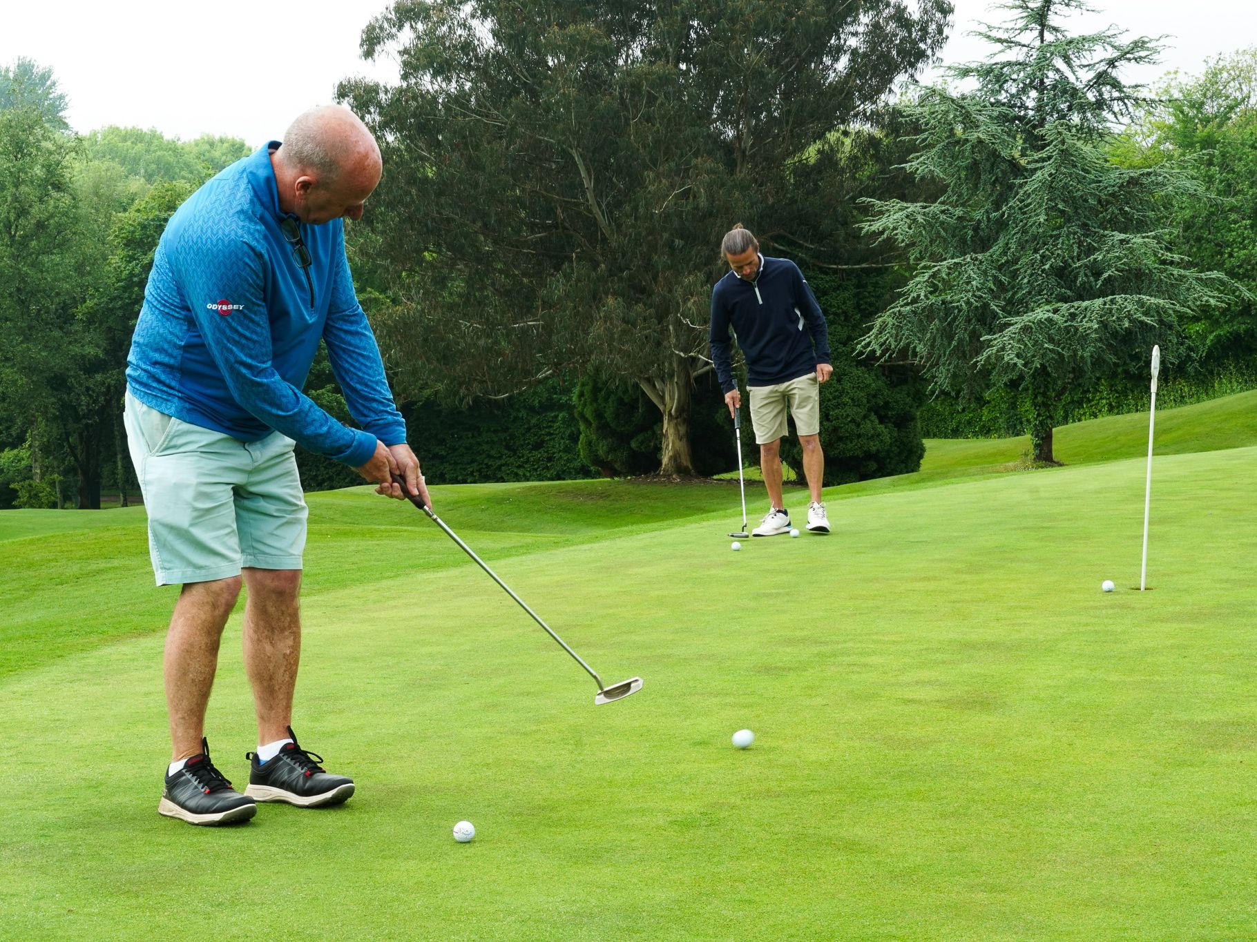 Two men playing golf on a putting green, both preparing to putt golf balls towards the hole, surrounded by trees and greenery.