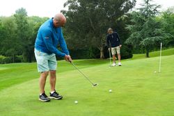 Two men playing golf on a putting green, both preparing to putt golf balls towards the hole, surrounded by trees and greenery.