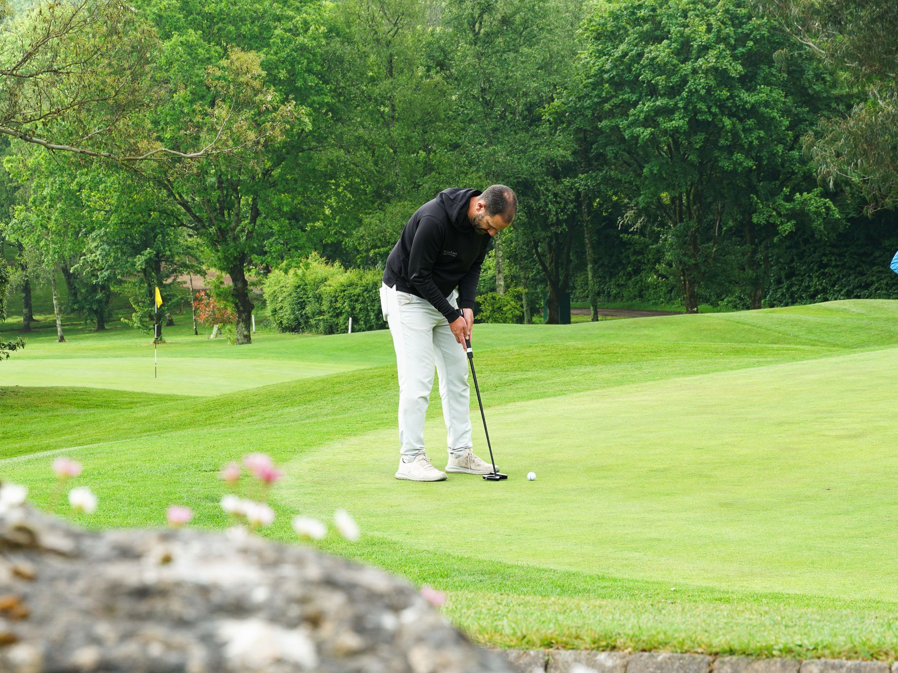 A man wearing a black hoodie and white pants is putting on a golf green surrounded by lush green trees.