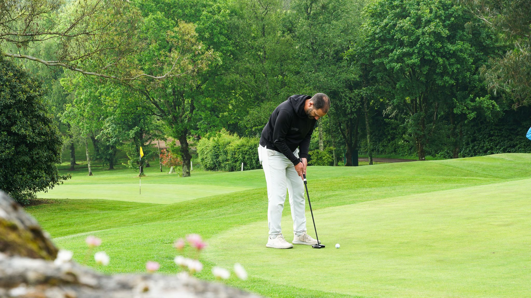A man wearing a black hoodie and white pants is putting on a golf green surrounded by lush green trees.