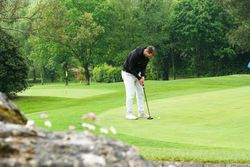 A man wearing a black hoodie and white pants is putting on a golf green surrounded by lush green trees.