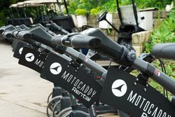 Close-up view of several Motocaddy rental electric golf trolleys lined up in front of parked golf carts outdoors.