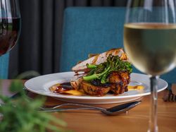 A plated meal featuring roasted meat, potatoes, and green vegetables, garnished with sauce, served on a white plate alongside glasses of red and white wine.