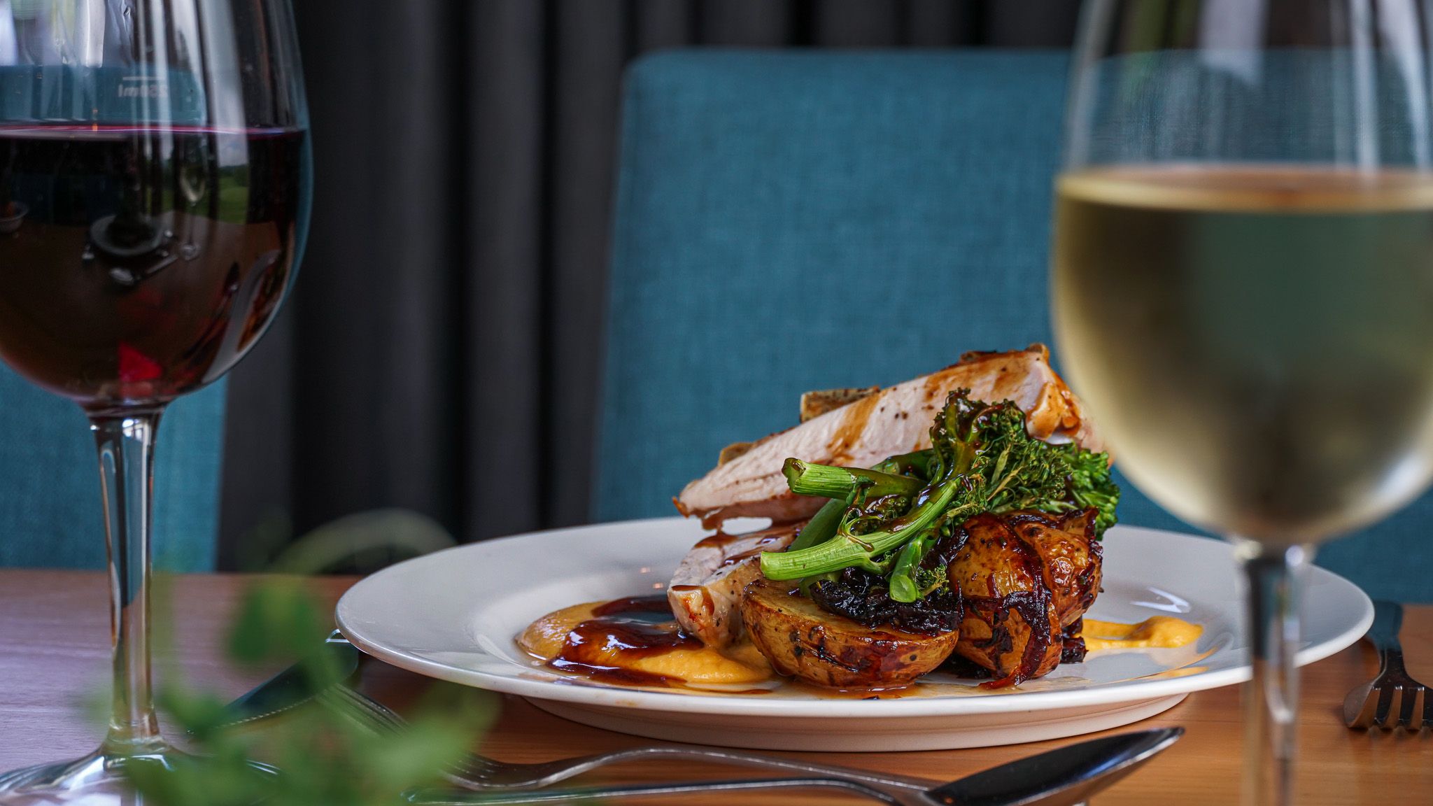 A plated meal featuring roasted meat, potatoes, and green vegetables, garnished with sauce, served on a white plate alongside glasses of red and white wine.