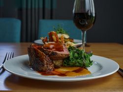 A gourmet plated meal featuring sliced roast meat with gravy, sautéed greens, a vegetable puree, and crispy garnish, served on a white plate with a glass of red wine on a wooden table.