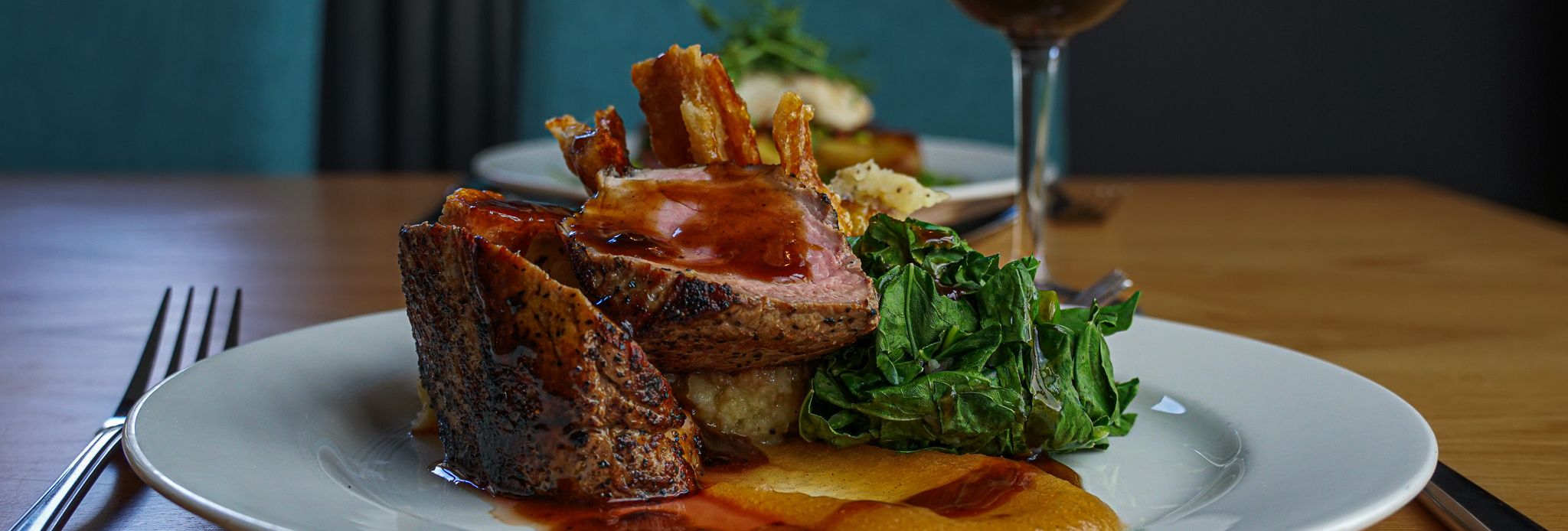 A gourmet plated meal featuring sliced roast meat with gravy, sautéed greens, a vegetable puree, and crispy garnish, served on a white plate with a glass of red wine on a wooden table.