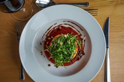 A gourmet salad with arugula, pine nuts, and balsamic drizzle served on a white plate, accompanied by a fork, knife, and spoon on a wooden table.