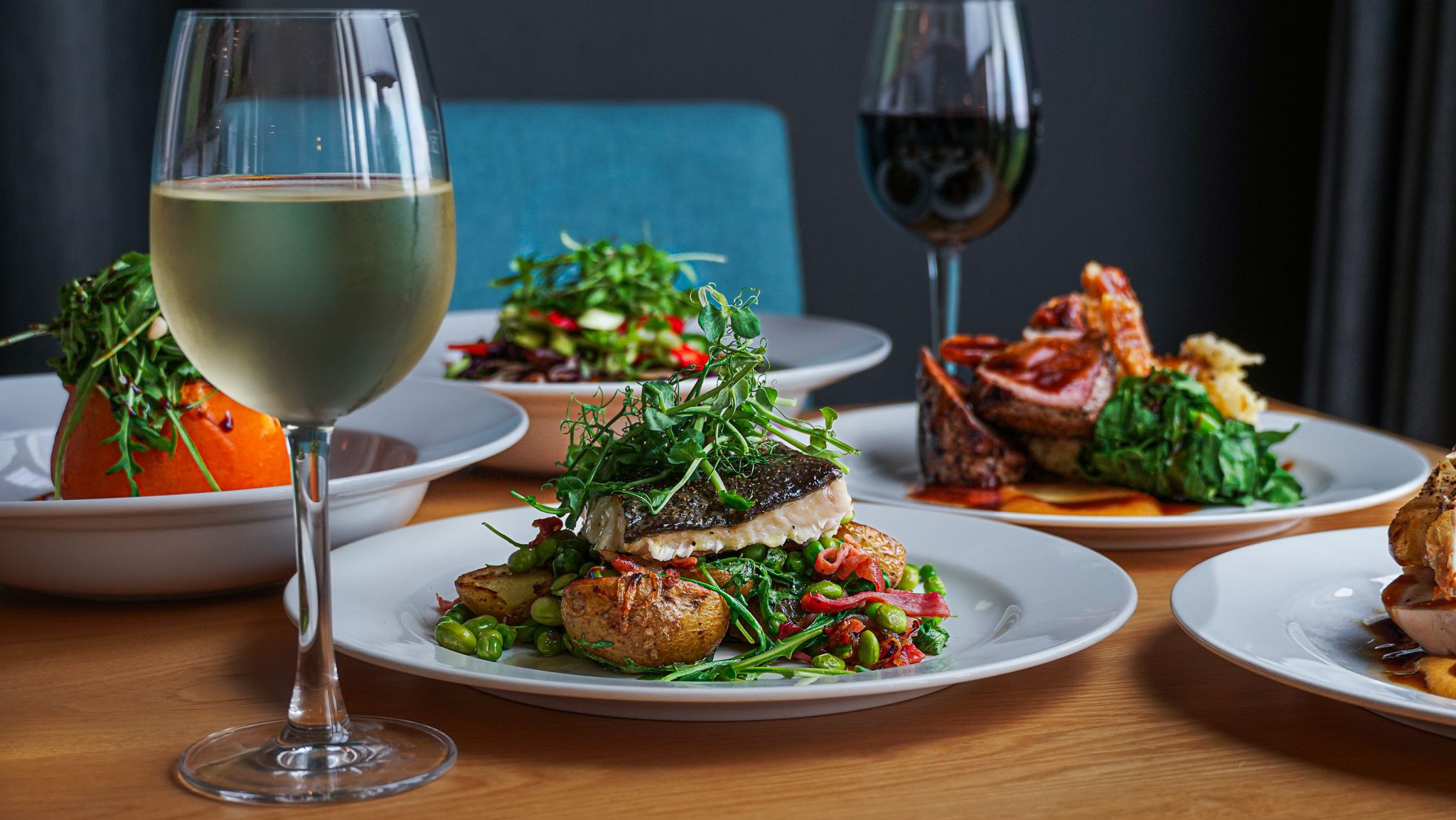 Fine dining dishes served on a table with a glass of white wine and a glass of red wine, featuring fish, vegetables, and gourmet sides.