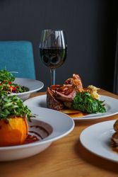 Plated gourmet meal with cooked meat, greens, mashed potatoes, and sauce, accompanied by a glass of red wine and other dishes on a wooden table.