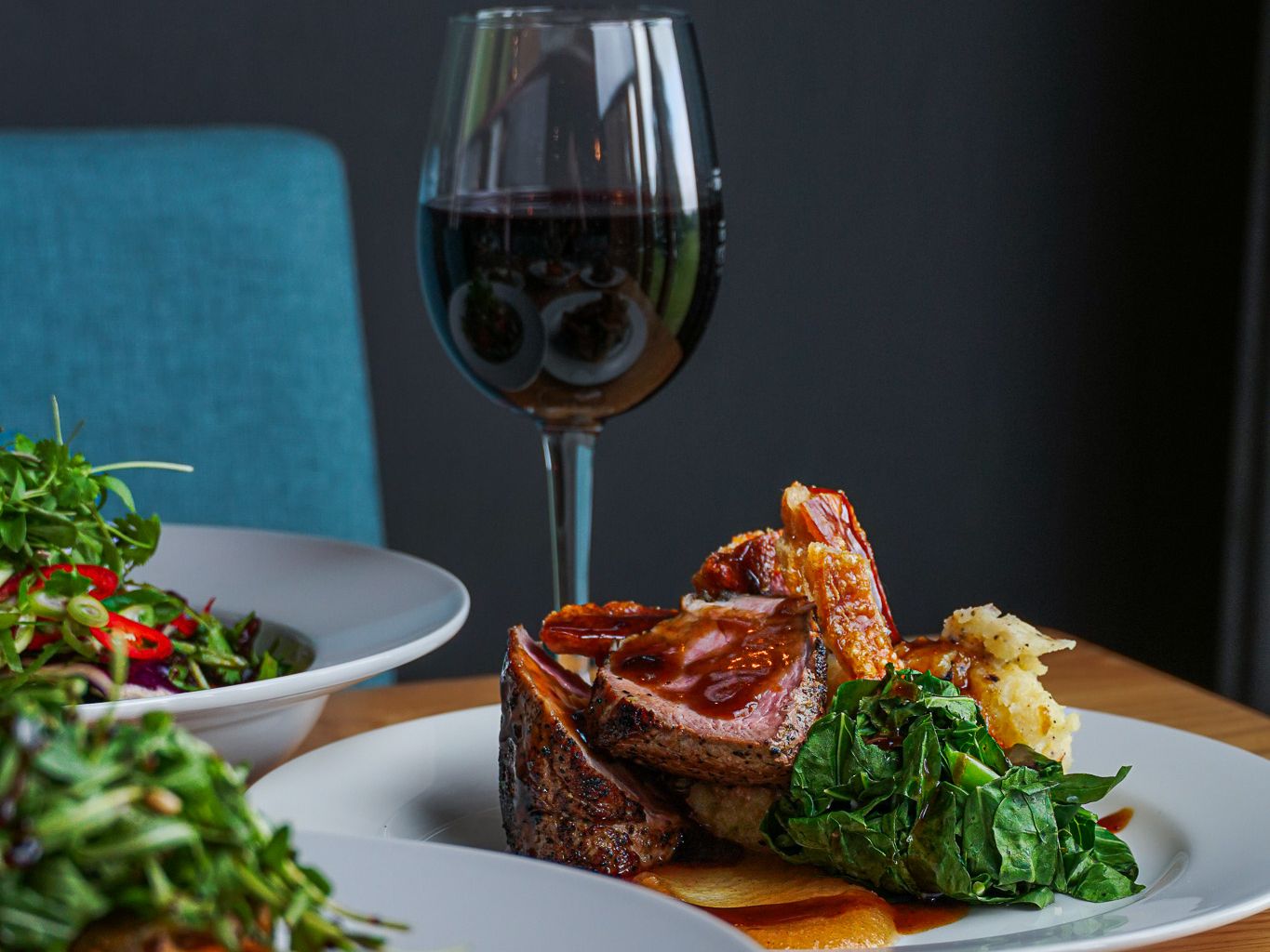 Plated gourmet meal with cooked meat, greens, mashed potatoes, and sauce, accompanied by a glass of red wine and other dishes on a wooden table.