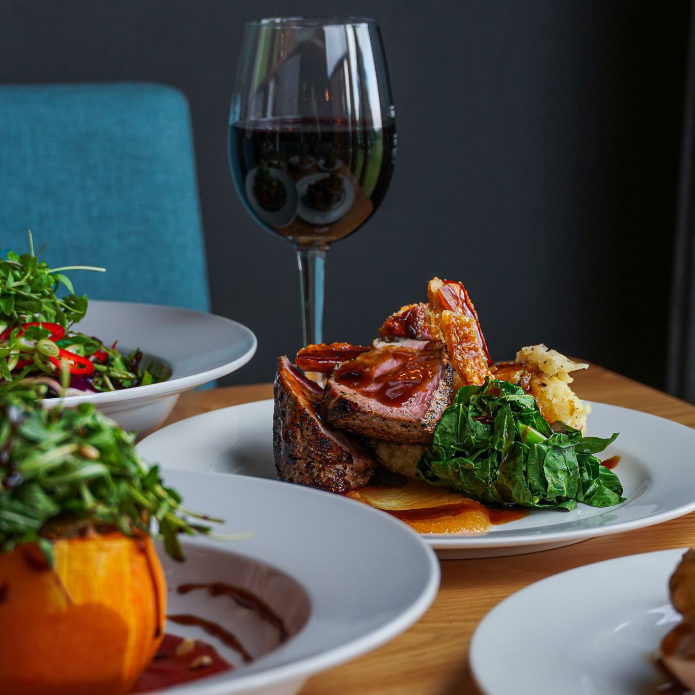 Plated gourmet meal with cooked meat, greens, mashed potatoes, and sauce, accompanied by a glass of red wine and other dishes on a wooden table.