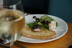 A plate with a slice of toasted bread, a piece of fish topped with greens, and a rolled cucumber appetizer filled with a creamy mixture, served next to a glass of white wine.