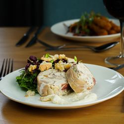 A plated meal featuring sliced stuffed chicken roulade, leafy greens salad with croutons, and a dollop of creamy sauce, set on a wooden table with cutlery and a glass of red wine.