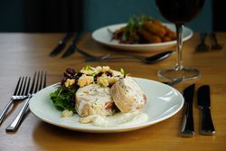 A plated meal featuring sliced stuffed chicken roulade, leafy greens salad with croutons, and a dollop of creamy sauce, set on a wooden table with cutlery and a glass of red wine.