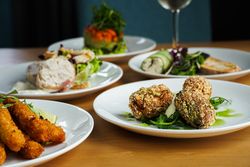 A selection of gourmet appetizers on white plates, including breaded and fried sticks, croquettes served on greens with sauce, and assorted vegetable and meat dishes, set on a wooden table.