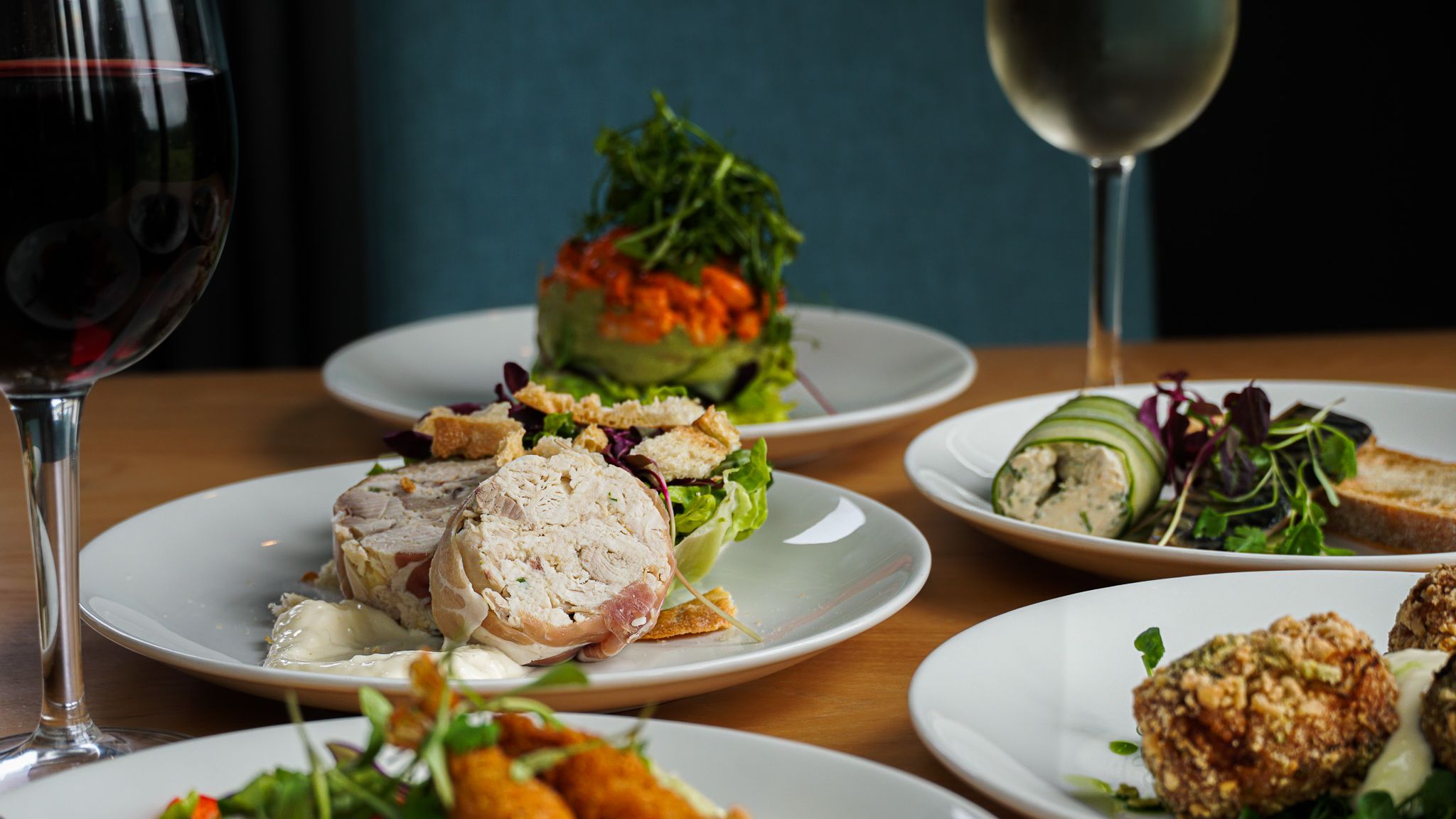 Assorted gourmet appetizers on white plates accompanied by glasses of red and white wine on a wooden table.