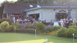 People dining outdoors at a restaurant next to a putting green, decorated with string lights and bunting, surrounded by colorful flowers and greenery.