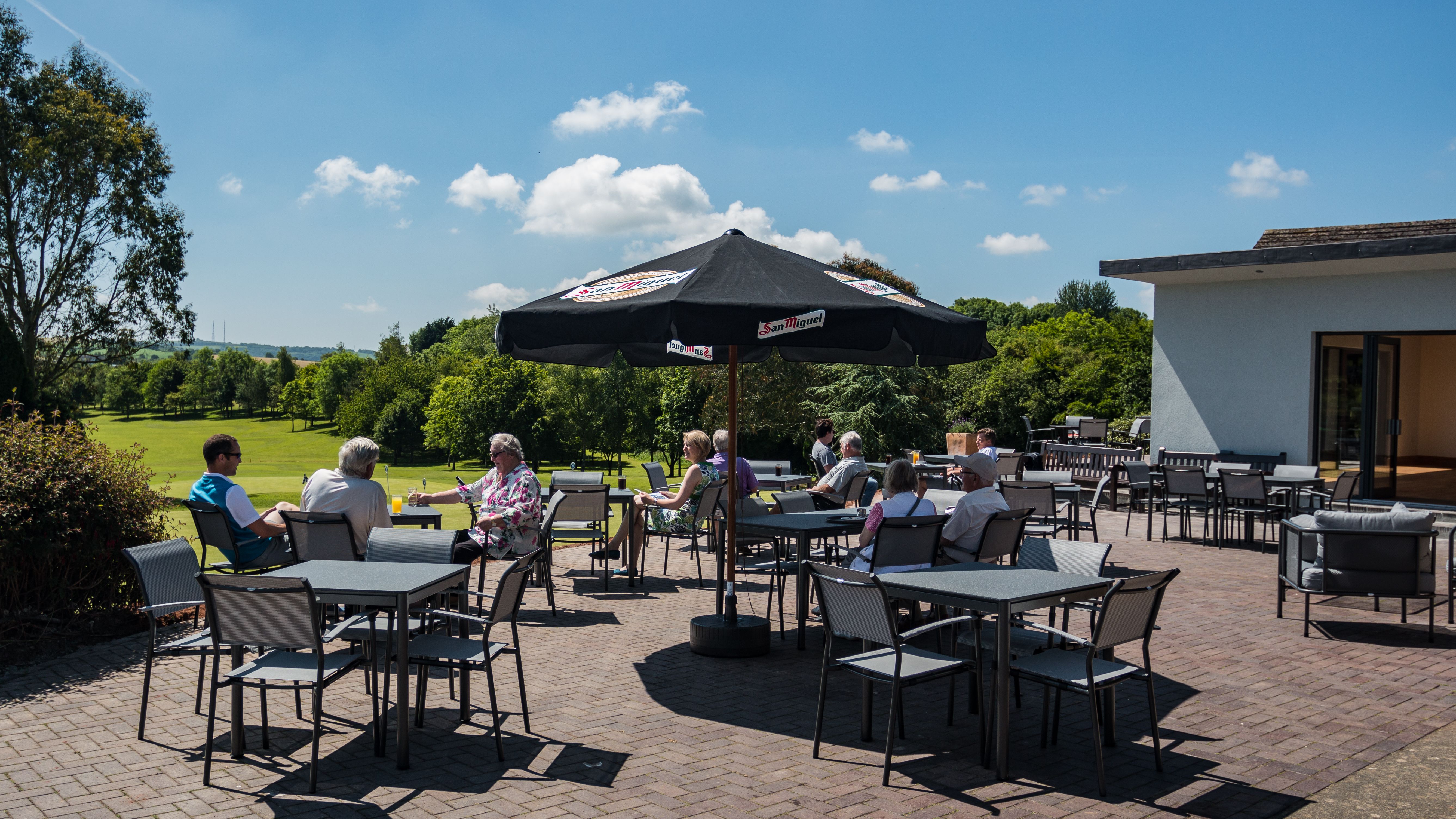 People sitting at outdoor terrace tables under a large umbrella on a sunny day, surrounded by greenery.