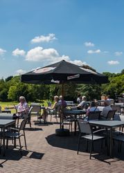 People sitting at outdoor terrace tables under a large umbrella on a sunny day, surrounded by greenery.