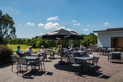 People sitting at outdoor terrace tables under a large umbrella on a sunny day, surrounded by greenery.