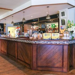 Interior of a cozy pub or bar with a wooden counter, drinks on display, and a menu written on a chalkboard wall.