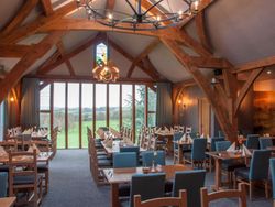 Spacious restaurant with wooden beams, blue chairs, large windows overlooking green fields, and neatly set tables.