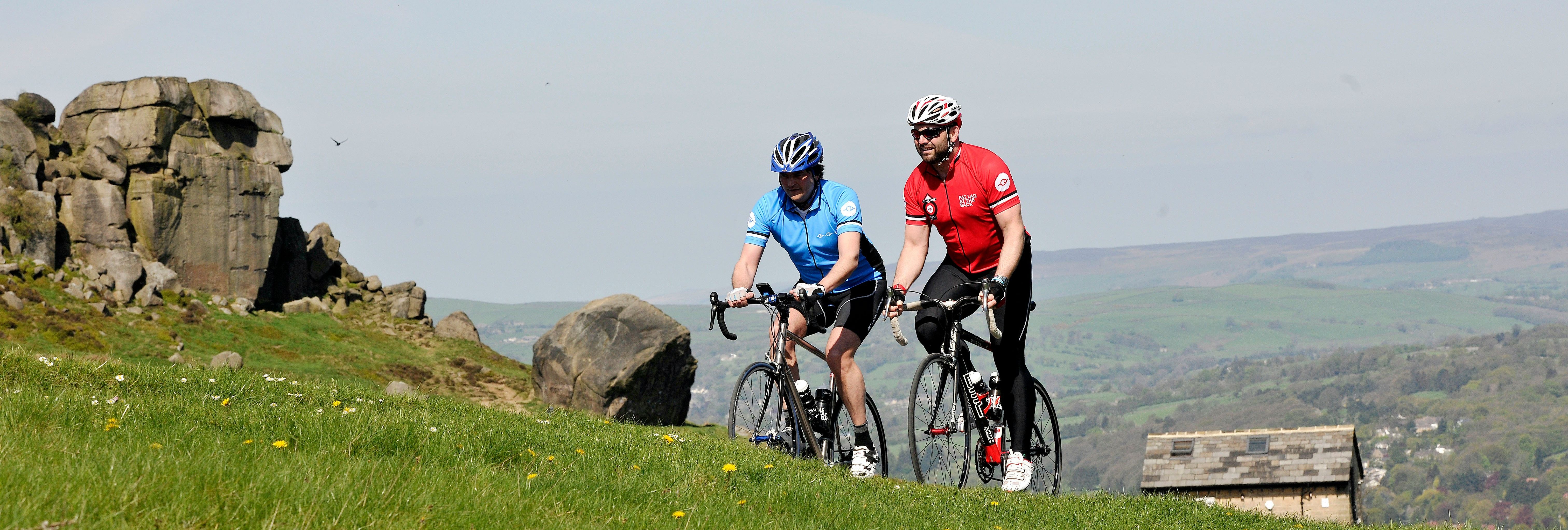 Two cyclists riding on a grassy hill with rocky outcrops and countryside in the background.
