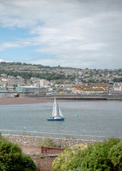 A small sailboat on a calm body of water with a town and hills in the background.