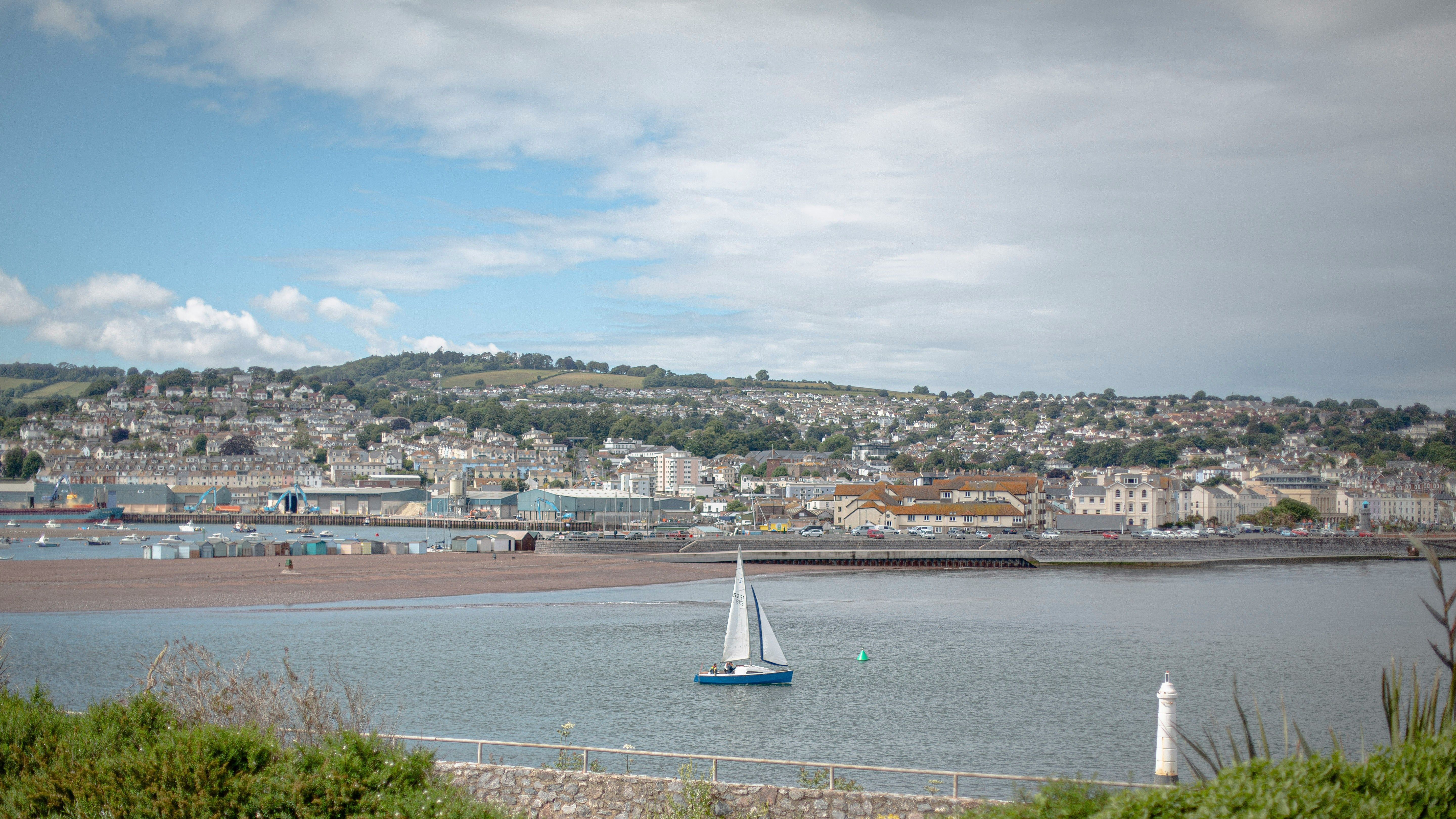 A small sailboat on a calm body of water with a town and hills in the background.
