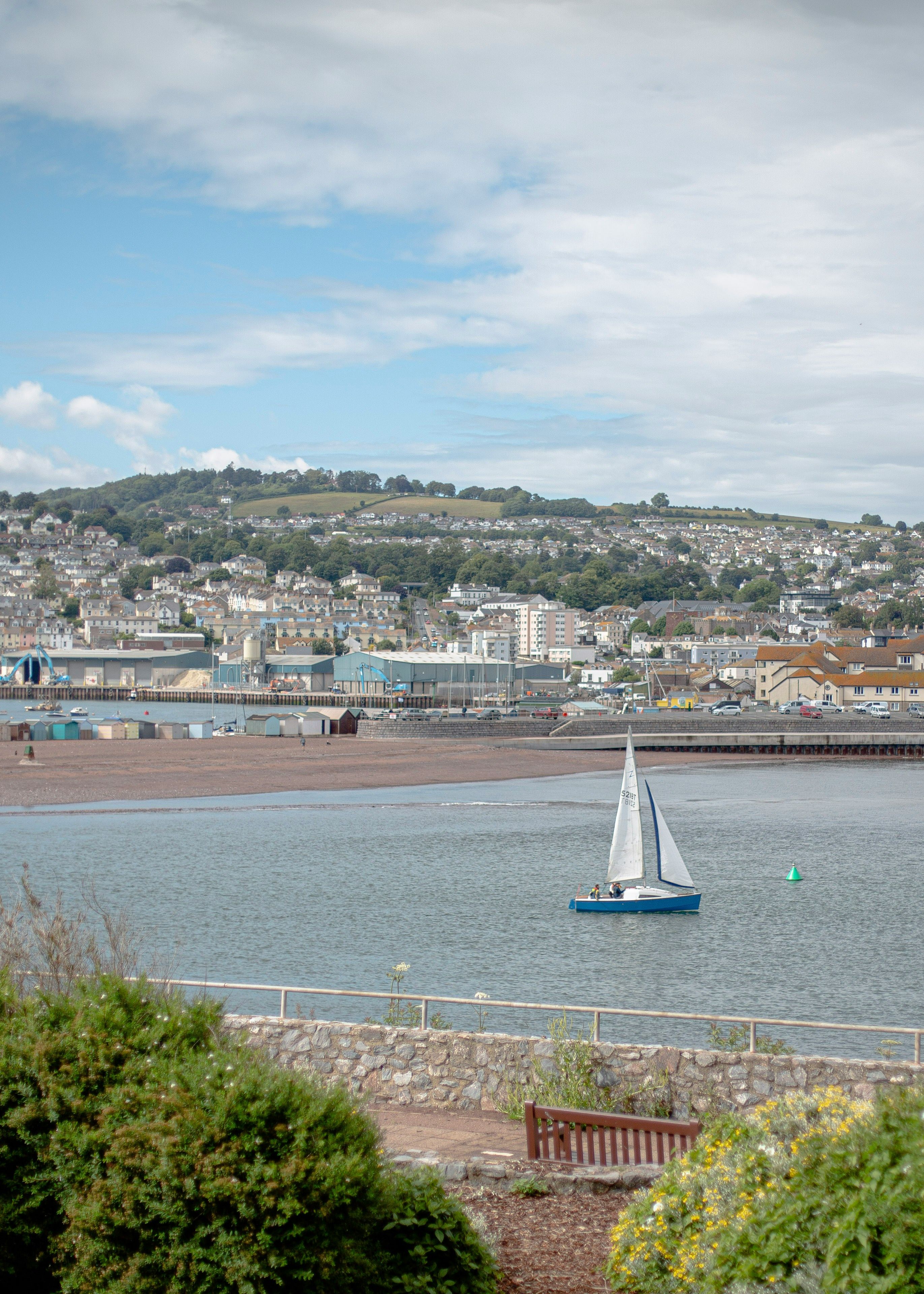 A small sailboat on a calm body of water with a town and hills in the background.