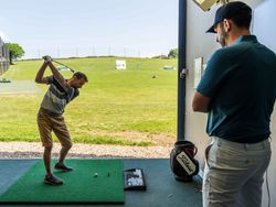 A man practices his golf swing at an indoor driving range as another man observes.