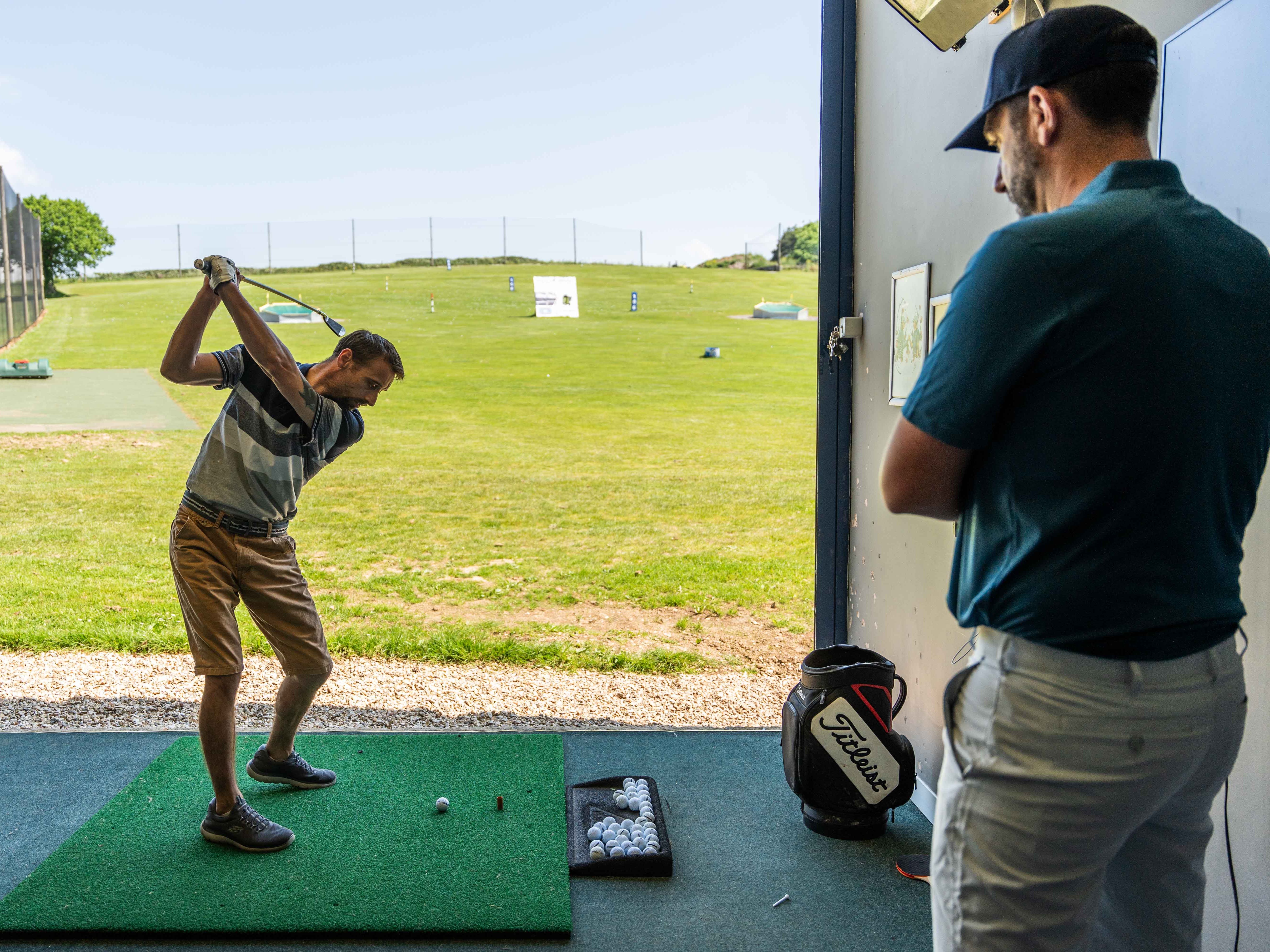 A man practices his golf swing at an indoor driving range as another man observes.