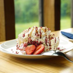 A plate of dessert with whipped cream, strawberries, and berry drizzle next to a glass of trifle dessert topped with strawberries