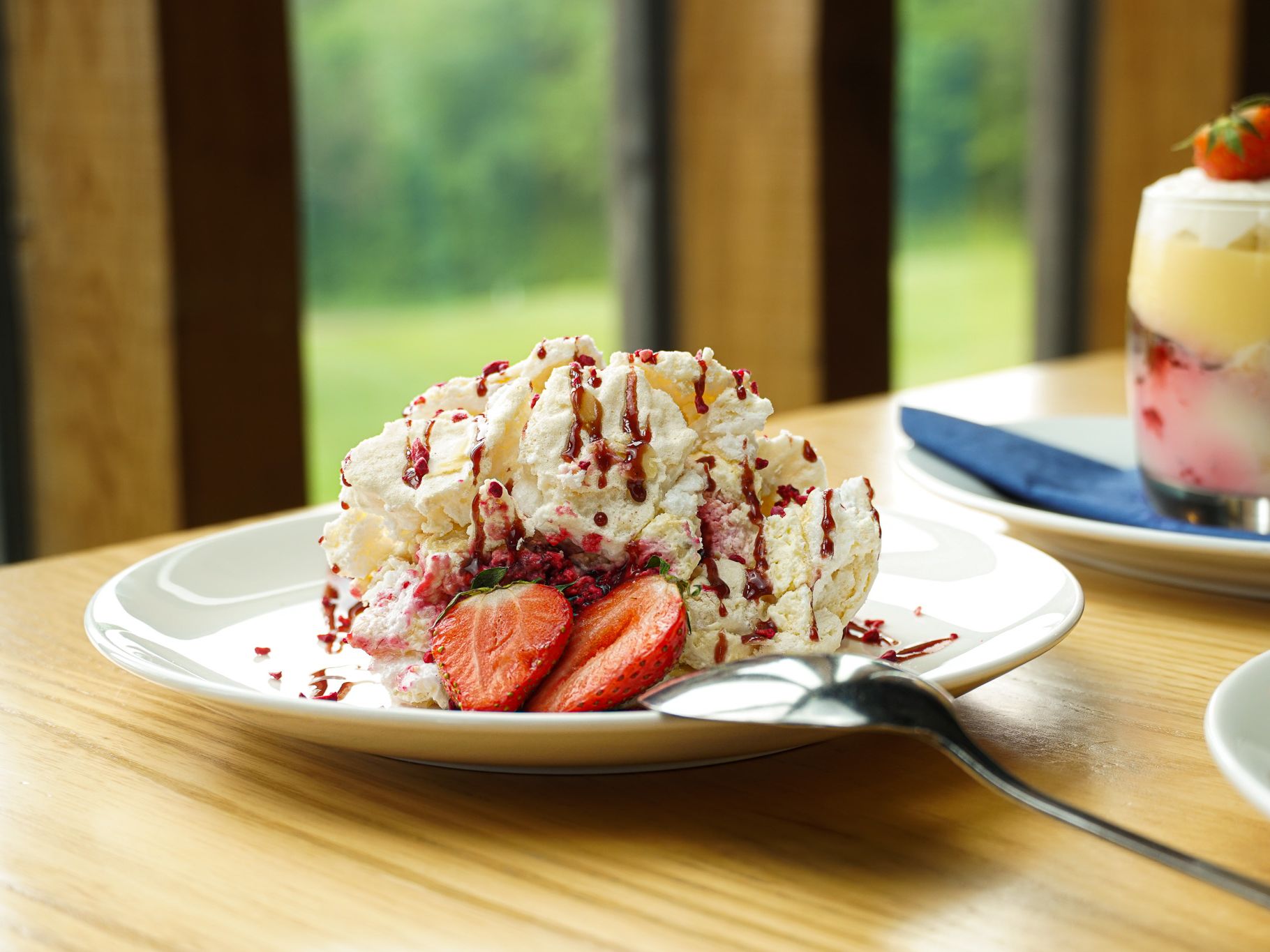 A plate of dessert with whipped cream, strawberries, and berry drizzle next to a glass of trifle dessert topped with strawberries
