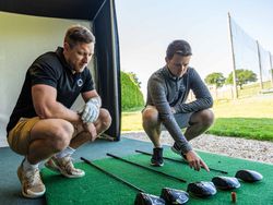 Two men squatting and examining several golf clubs laid out on a mat at a driving range.