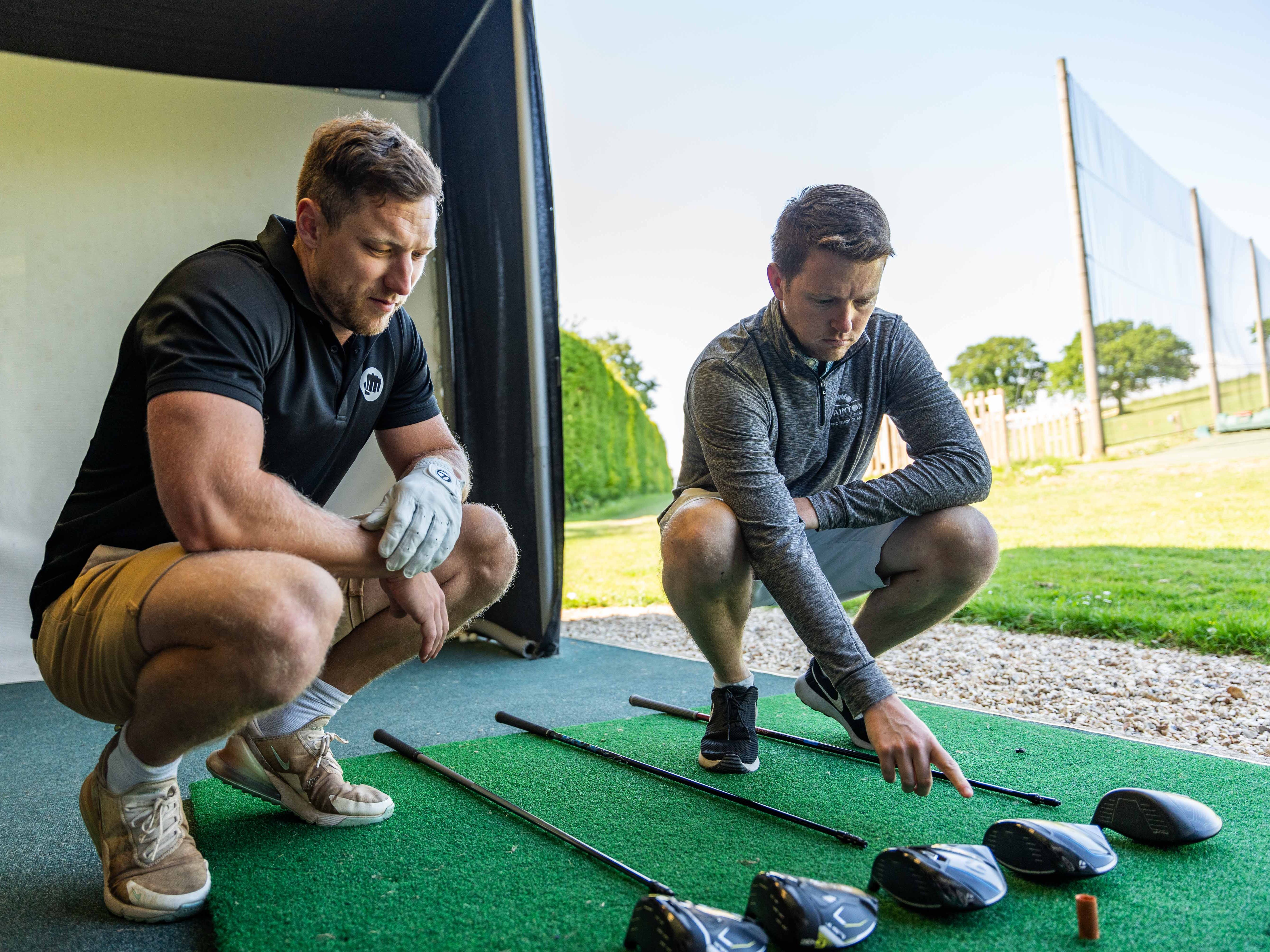 Two men squatting and examining several golf clubs laid out on a mat at a driving range.