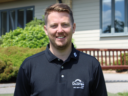 Man in a black polo shirt standing outdoors in front of a building with greenery and a bench.