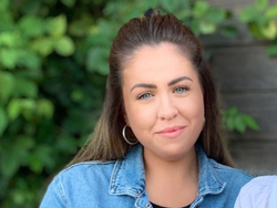 Woman with long brown hair wearing a denim jacket, sitting outdoors.