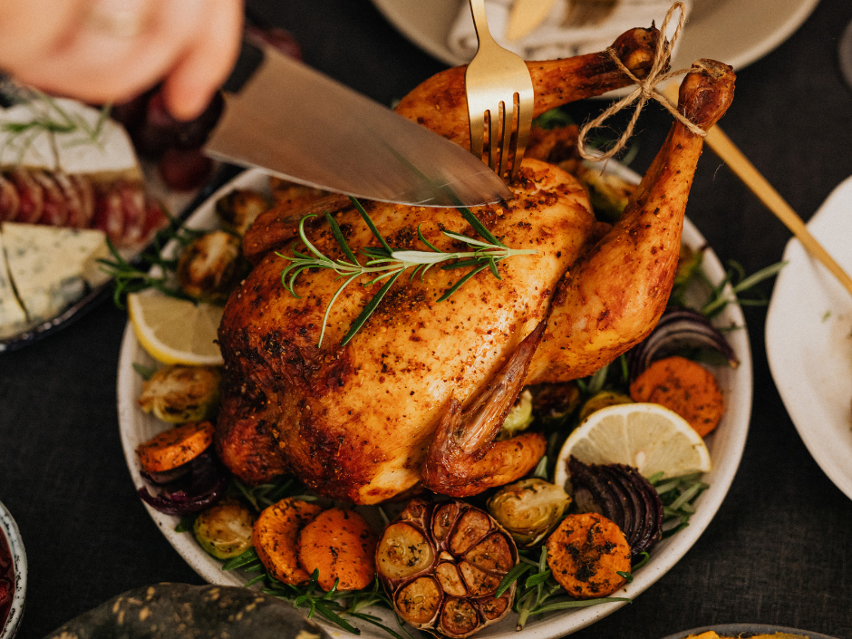Person carving a roasted chicken garnished with herbs, lemons, and roasted vegetables.