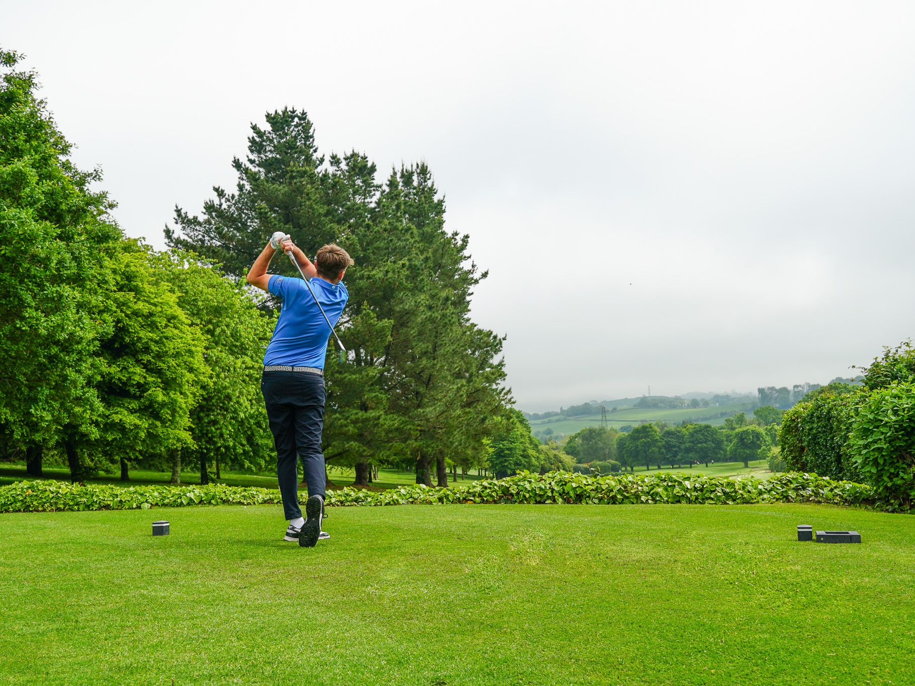 Golfer swinging club on a lush green golf course with trees and hills in the distance
