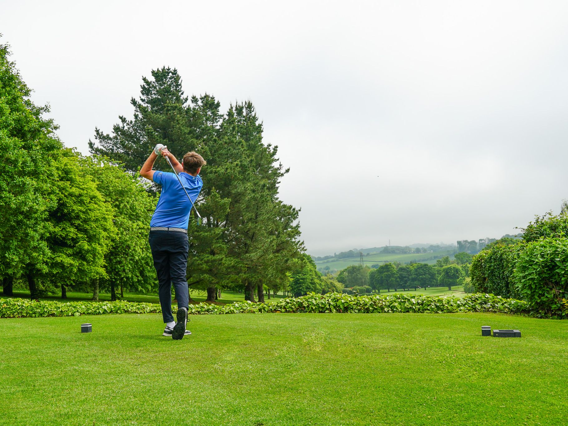 Golfer swinging club on a lush green golf course with trees and hills in the distance