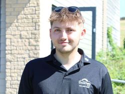 A young man wearing a black collared shirt with a logo and sunglasses on his head, standing outside in front of a building.