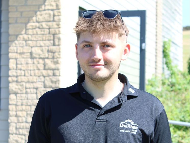A young man wearing a black collared shirt with a logo and sunglasses on his head, standing outside in front of a building.