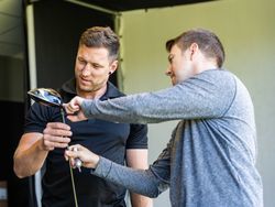 Two men inspecting a golf club indoors, one holding the club while the other points at it.