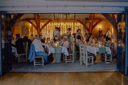 People seated and standing around decorated tables at an indoor event with fairy lights and wooden beams.
