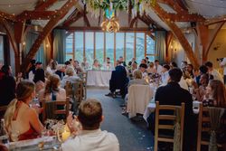 A wedding reception inside a rustic venue with wooden beams, where guests are seated at tables facing a bridal party at the head table.
