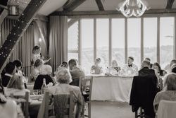 A black and white photograph of a wedding reception, showing a woman giving a speech to seated guests and the bridal party at the head table.